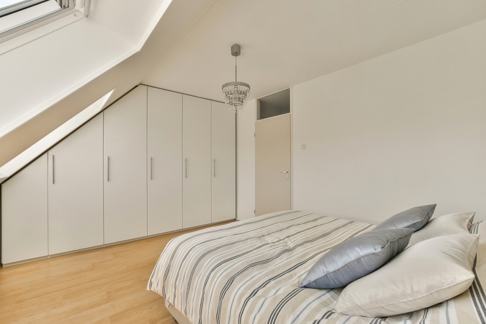 Chambre mansardée lumineuse avec placard blanc sur mesure, lit rayé et parquet clair sous un toit incliné.